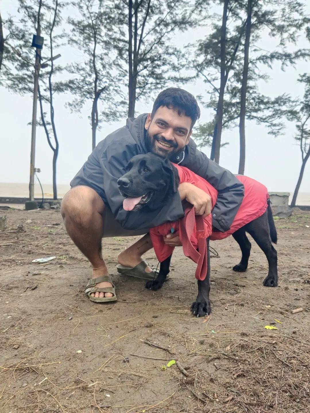 A pet parent crouching on a beach, hugging a black Labrador in a red raincoat